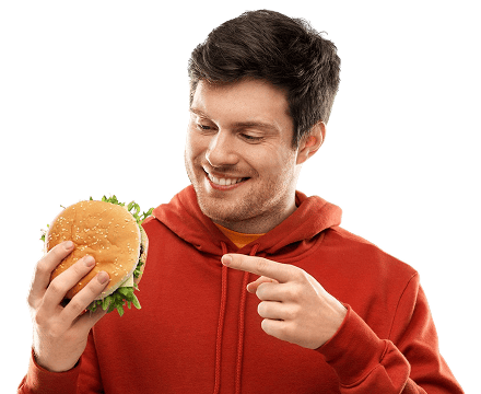 Açık renk bir arka plan önünde, elinde bir hamburger tutan ve onu işaret eden, gülümseyen yalnız bir adam.